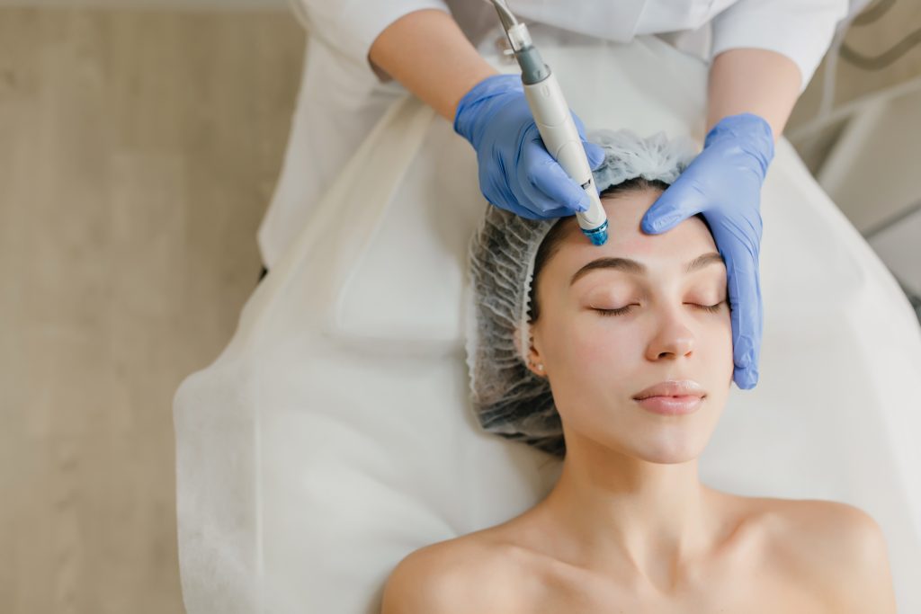 A woman lies with her eyes closed while a skincare professional wearing blue gloves uses a handheld cosmetic device on her forehead during a facial treatment.