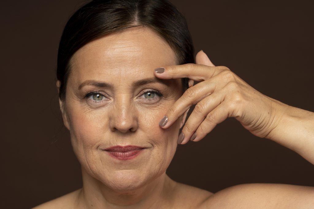 A close-up portrait of a middle-aged woman with light skin and green eyes, looking directly at the camera with a slight smile. She has dark brown hair pulled back and is wearing subtle makeup. One hand is raised to her face, with her fingers gently touching her temple and cheek. The background is a smooth, warm brown tone.
